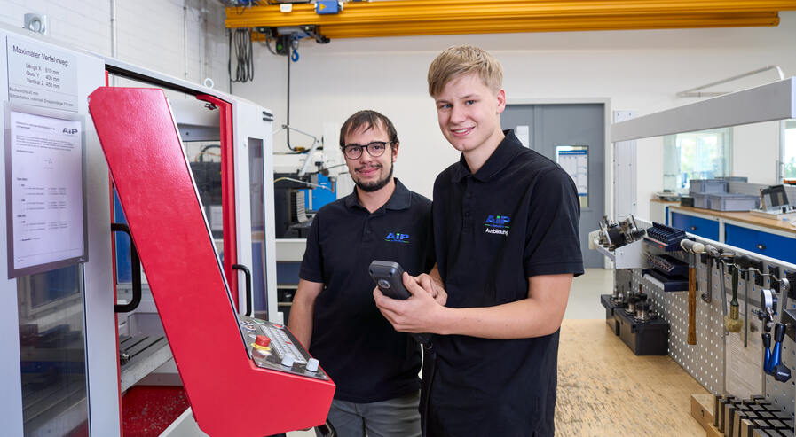 Ausbilder und Auszubildender stehen gemeinsam an der Fräsmaschine und lachen in die Kamera.