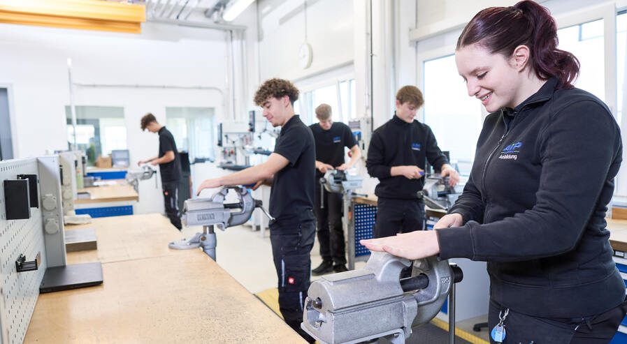 Fünf Auszubildende sind mit Feilen in der Lehrwerkstatt beschäftigt und wirken dabei glücklich.