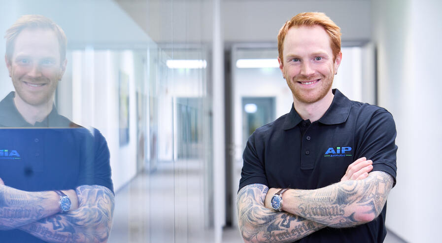 Mitarbeiter steht im Fokus des Bildes und lächelt mit verschränkten Armen in die Kamera.