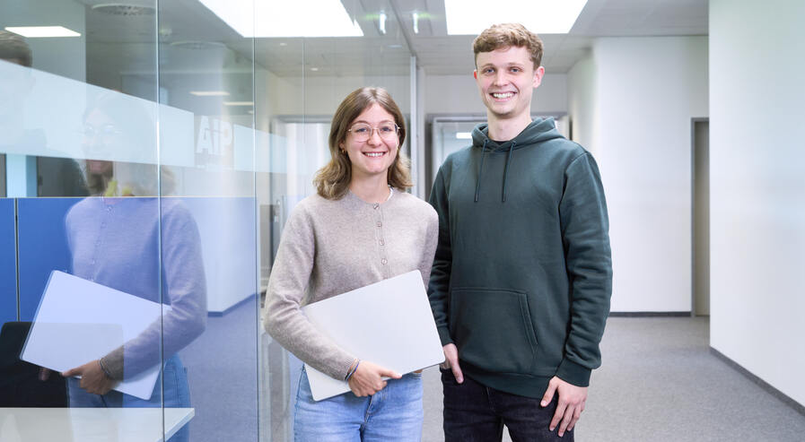 Zwei Studierende stehen nebeneinander und lächeln in die Kamera. Die Dame hält ihren Laptop unter dem Arm.